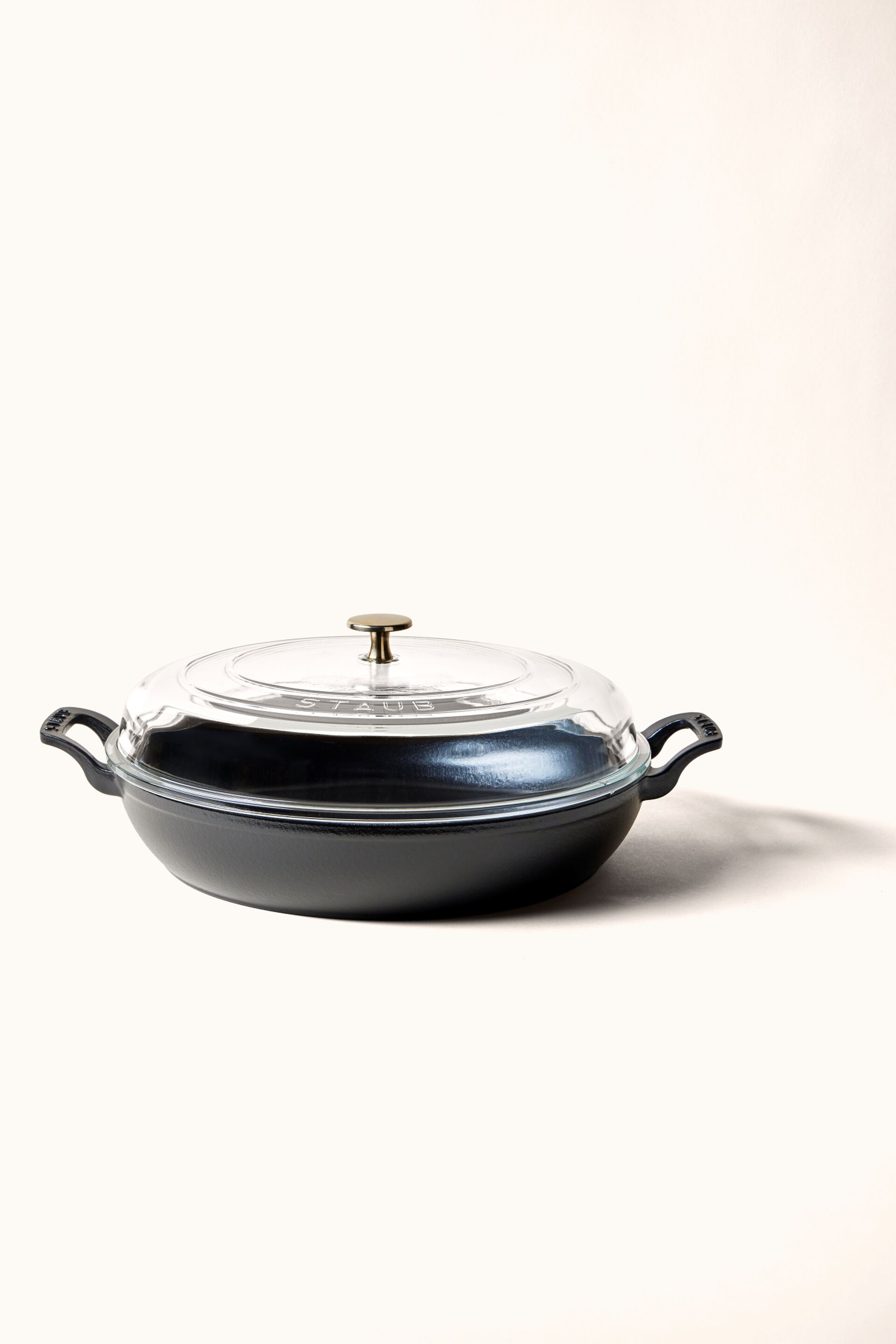 A light grey Staub braiser sits on a grey tablecloth atop a countertop, adjacent to a cutting board laden with bread and garlic to the side, with salt and pepper shakers nearby, creating a cozy and inviting kitchen scene.